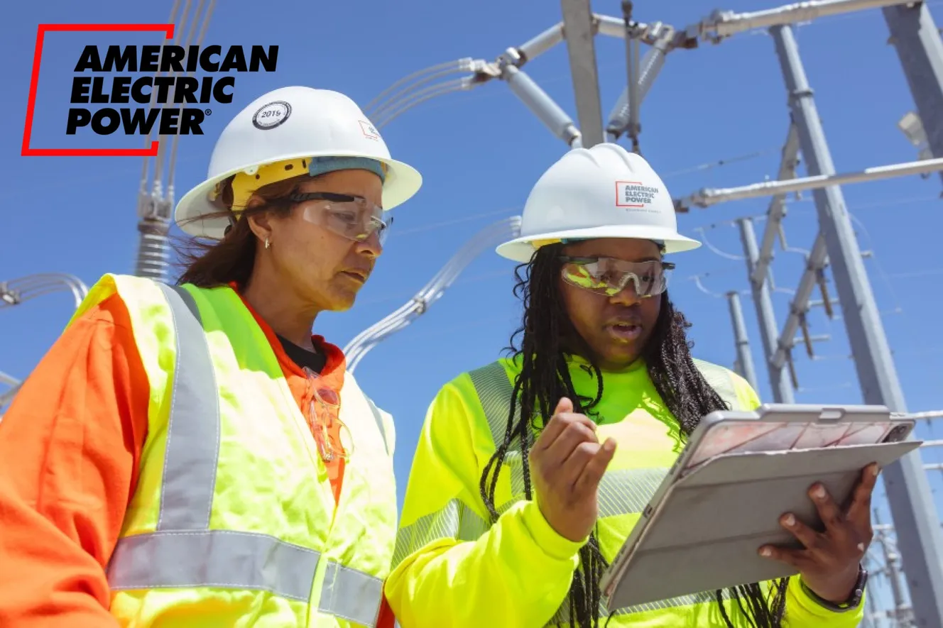 Two utility workers in hard hats and safety vests review data on a tablet at an American Electric Power substation.