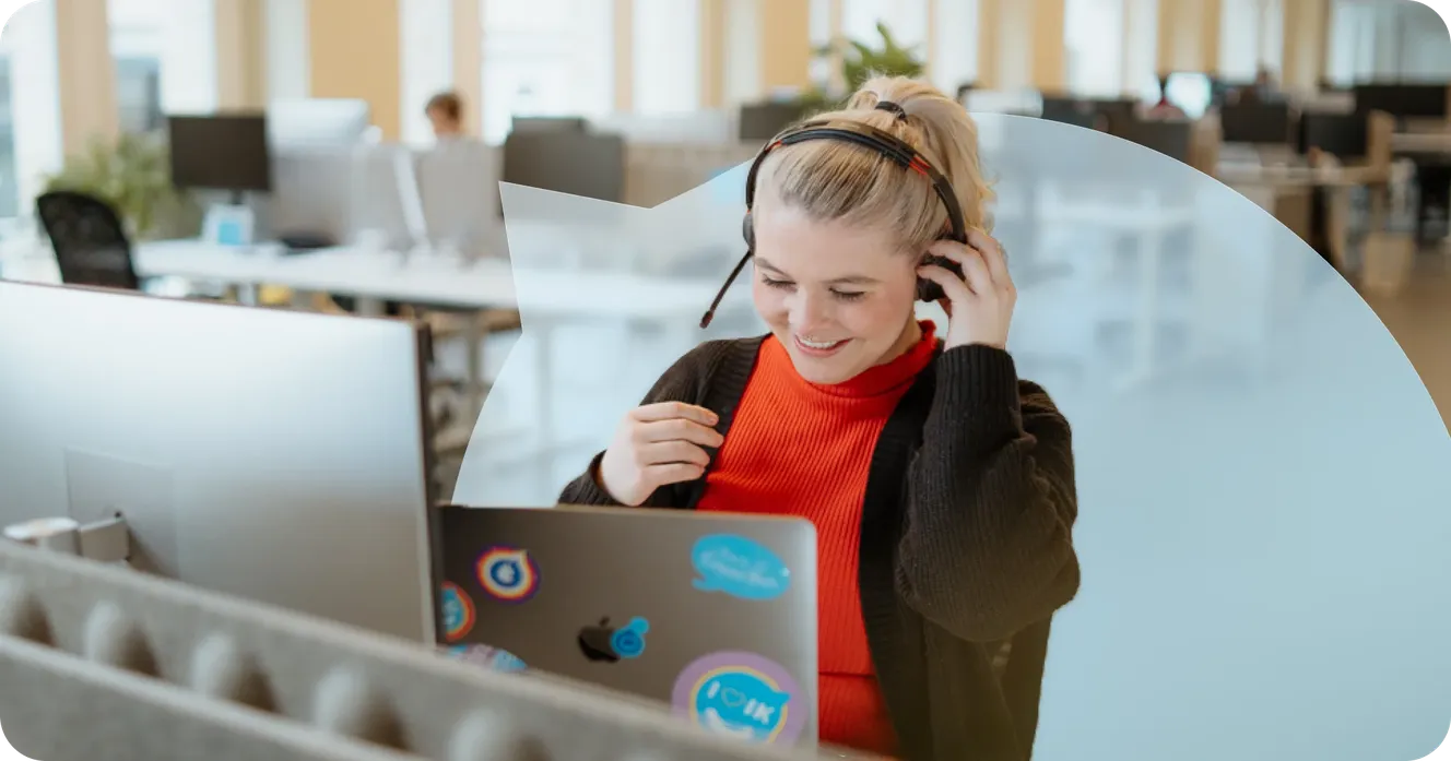 Staffbase employee smiling while putting on a headset at her laptop in an open-plan office.