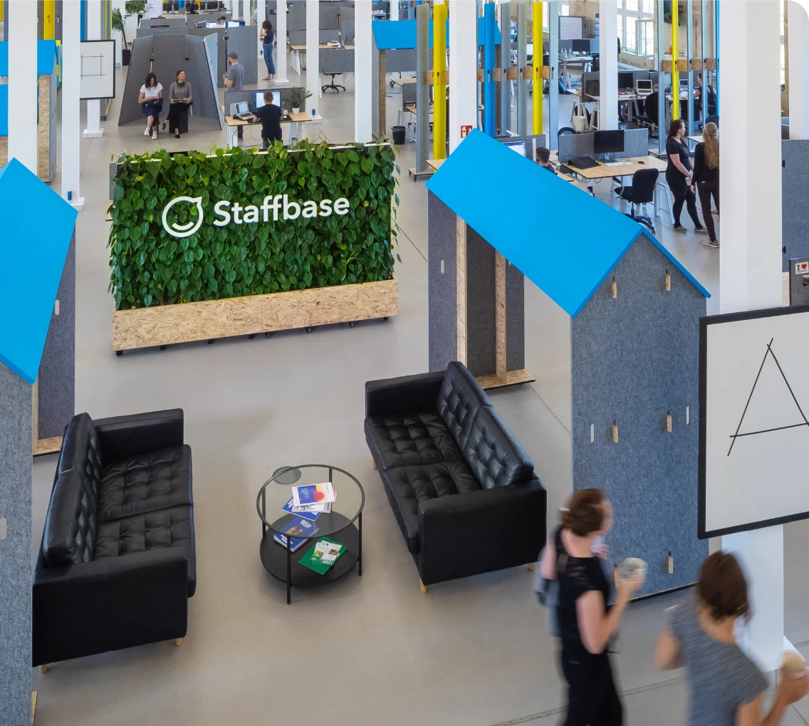 Modern office space with black sofas, a round glass table, and a green wall with the "Staffbase" logo. People walk by, and small structures are visible.