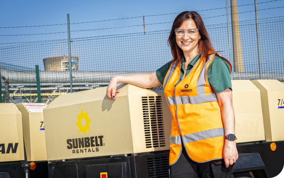Woman in an orange safety vest and glasses smiles, standing next to Sunbelt Rentals equipment, with industrial background.