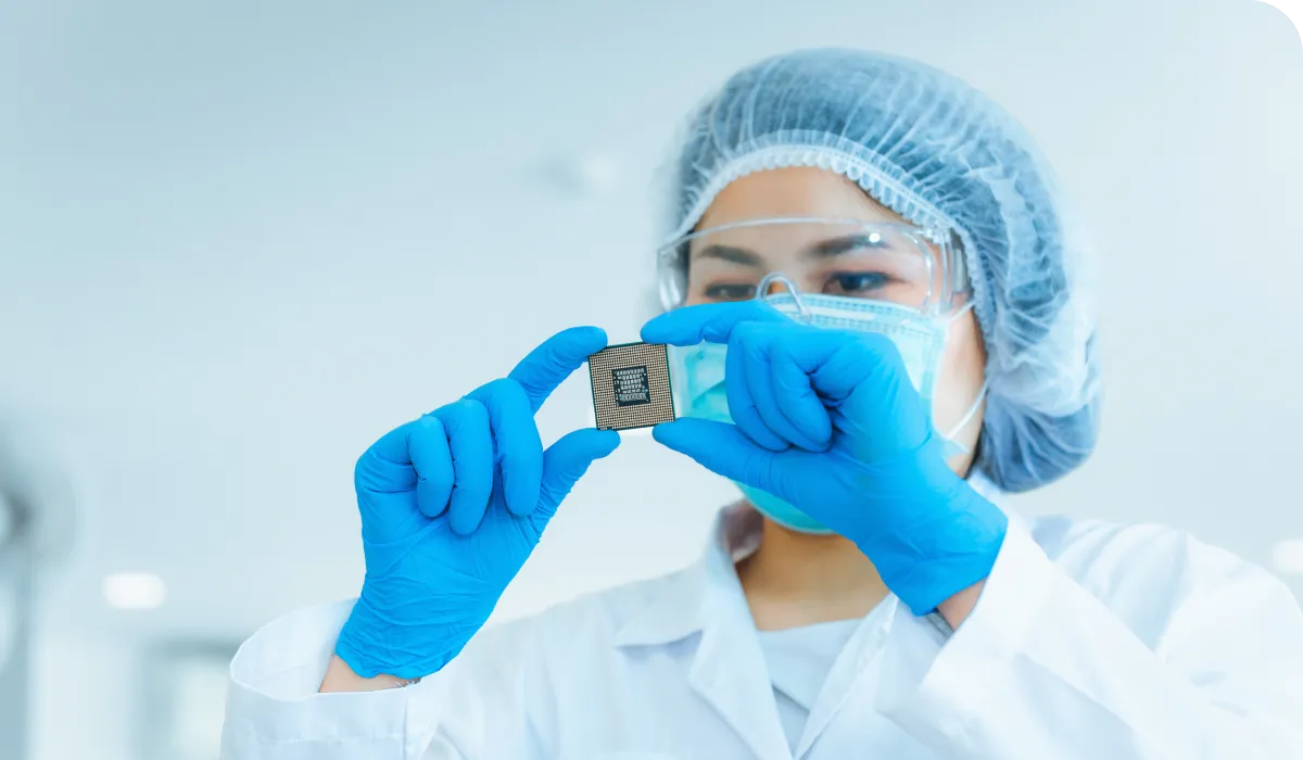 Scientist in protective gear examining a microchip, wearing a lab coat, hairnet, mask, and blue gloves, in a bright laboratory setting.