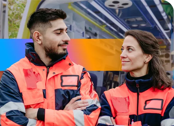 Two people in orange safety jackets stand side by side, smiling at each other, inside a brightly lit vehicle.