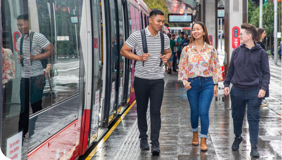 Three people walking on a wet tram platform, smiling and chatting. A tram is parked beside them.