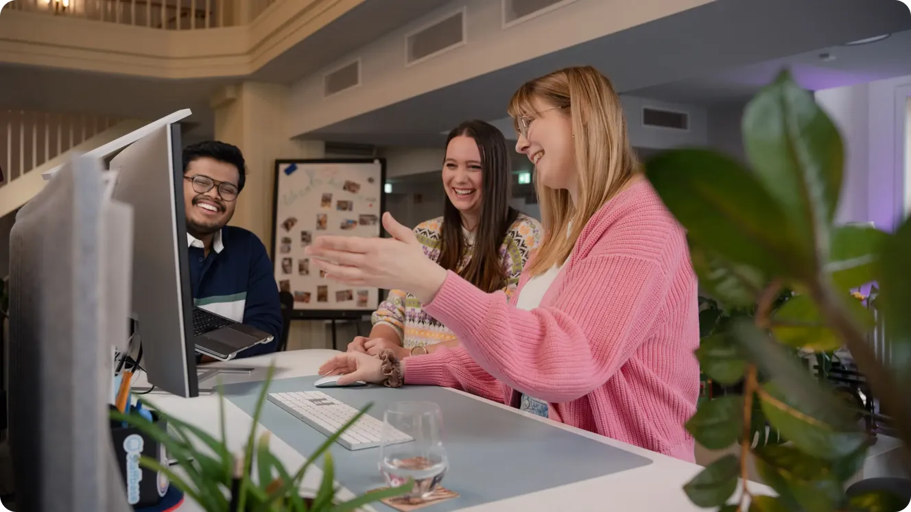 Three Staffbase colleagues laughing together while working at a desktop computer in a bright office atrium.