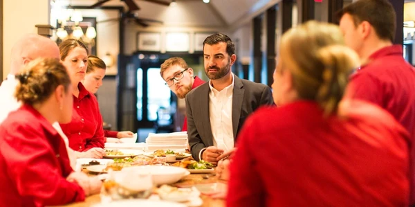 A group of restaurant staff in red shirts listens attentively to a man in a suit during a meeting at a wooden table with food dishes.