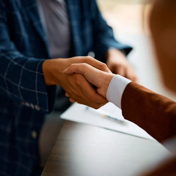 Two people in business attire shaking hands over a table with documents, symbolizing agreement or partnership.