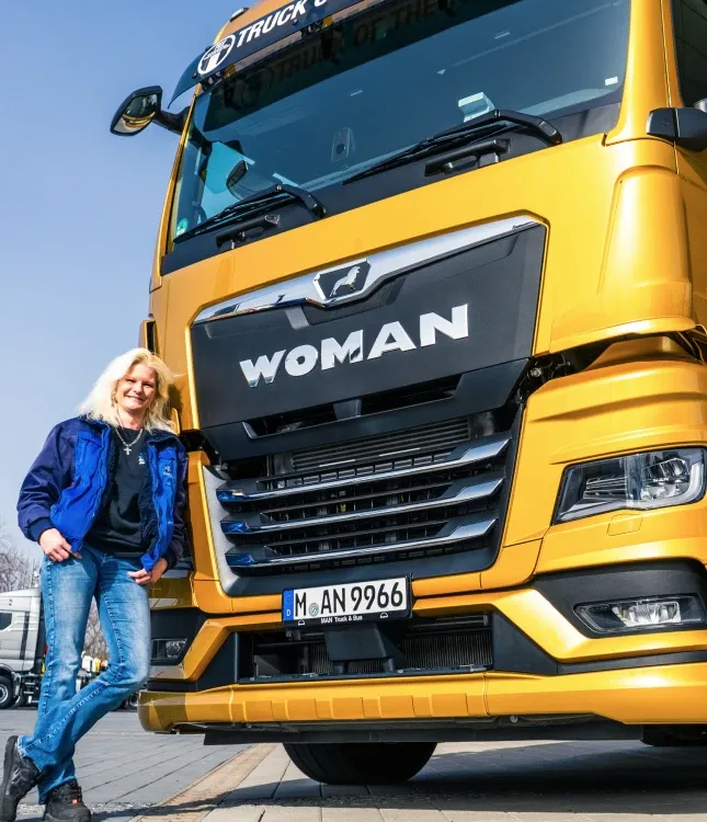 Person in a blue jacket leans against a large yellow truck with "WOMAN" on the front, parked outdoors on a sunny day.