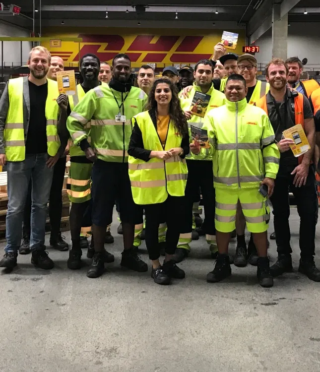 Group of DHL workers in high-visibility vests stand together in a warehouse, smiling and holding small packages.