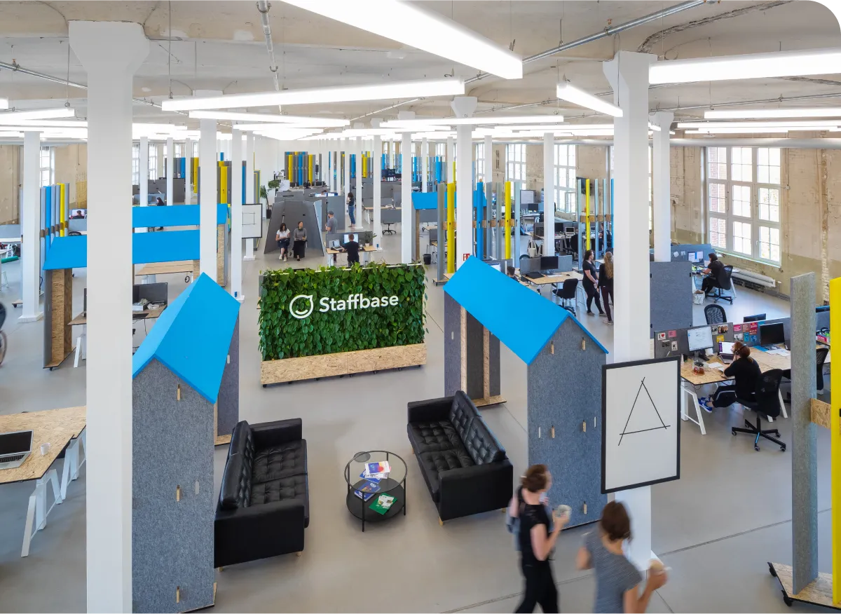 Spacious modern office with open workspaces, a central green wall with "Staffbase" logo, and people walking and working at desks.