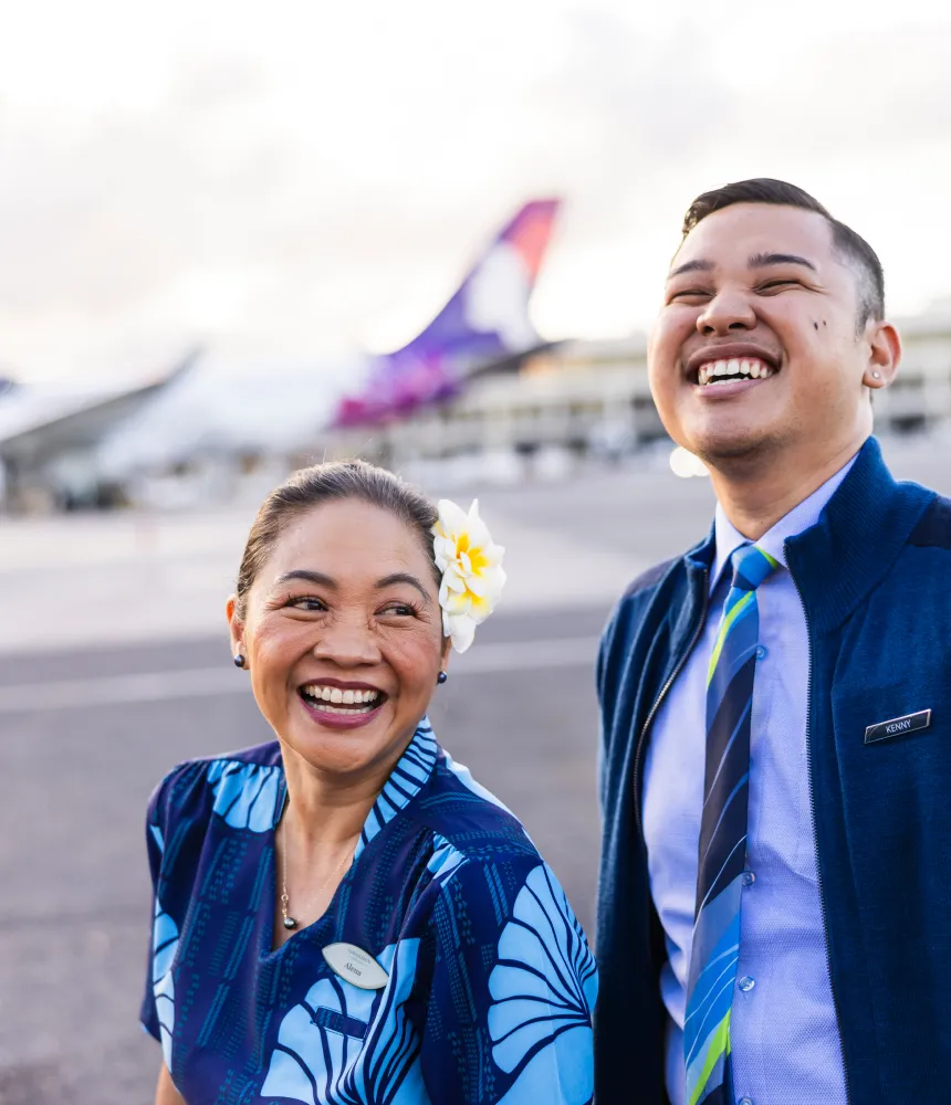 Two airline employees smiling on a runway, with one wearing a floral dress and the other in a uniform with a tie; airplane in the background.