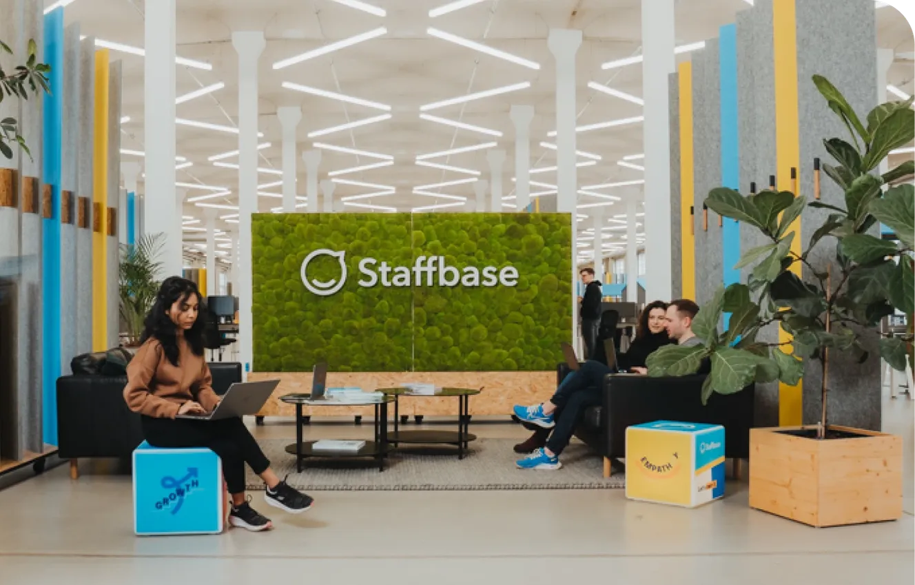 Modern office space with people sitting on sofas, working on laptops. A green wall displays the "Staffbase" logo. Bright, open environment.